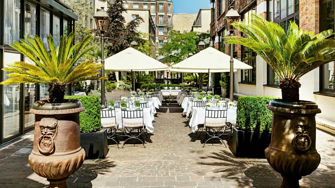 Utendørs selskapsområde med bord og parasoller omgitt av bygninger og palmer. (AI-generert tekst)