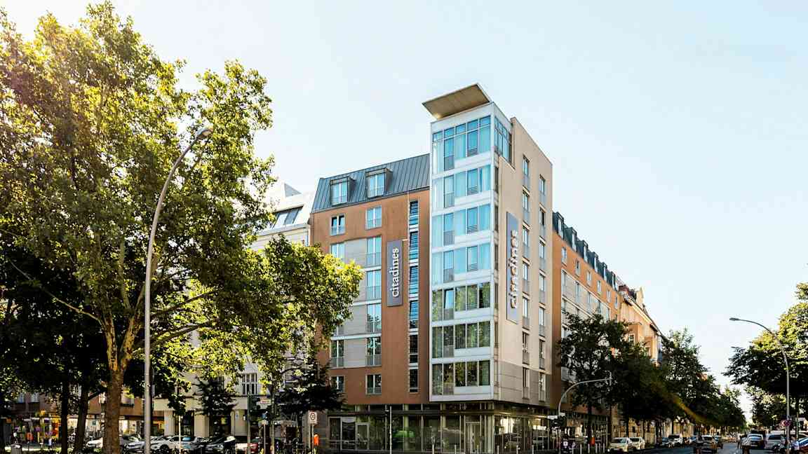 Et moderne hotell med glassfasade omgitt av trær og bymiljø. (AI-generert tekst)