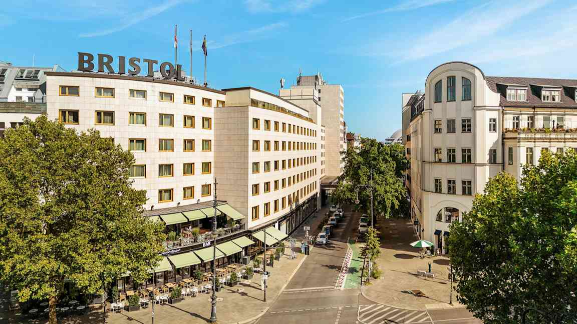 Et bylandskap som viser en bygning med teksten "Bristol" på taket, omgitt av trær og en gate med uteservering. (AI-generert tekst)