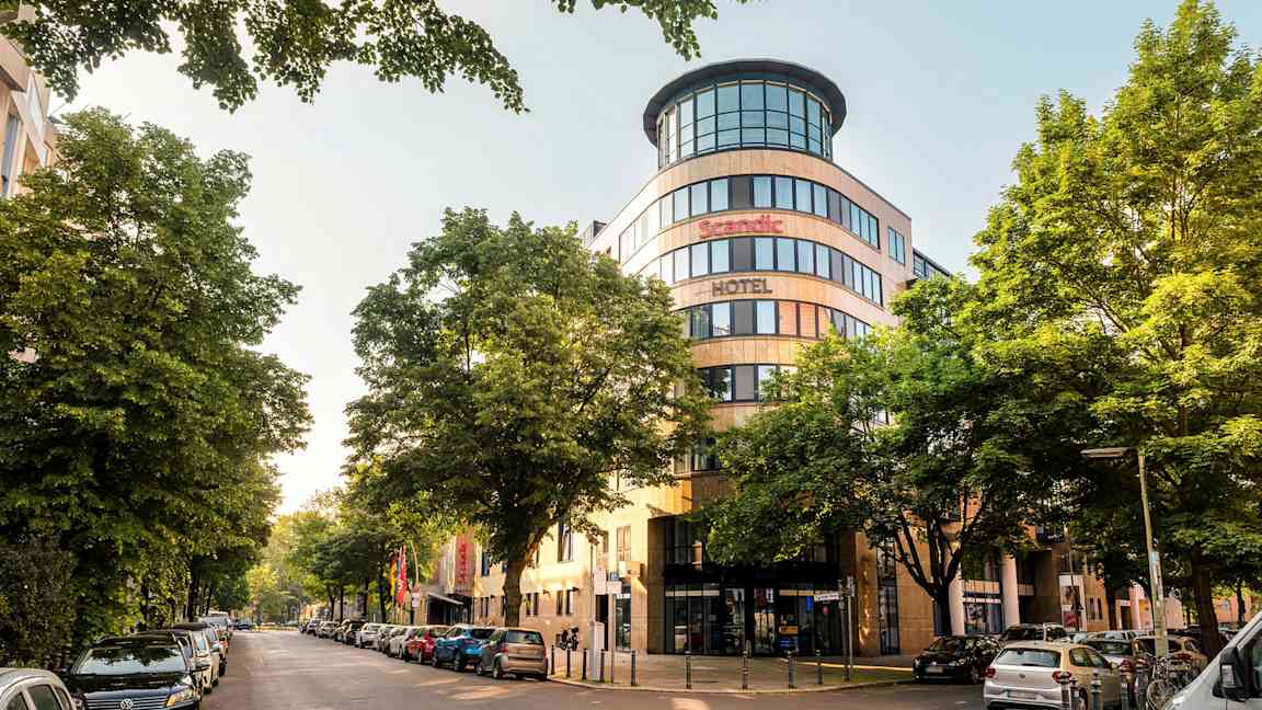 En byutsikt med fasaden til et Scandic-hotell omgitt av trær og parkerte biler. (AI-generert tekst)