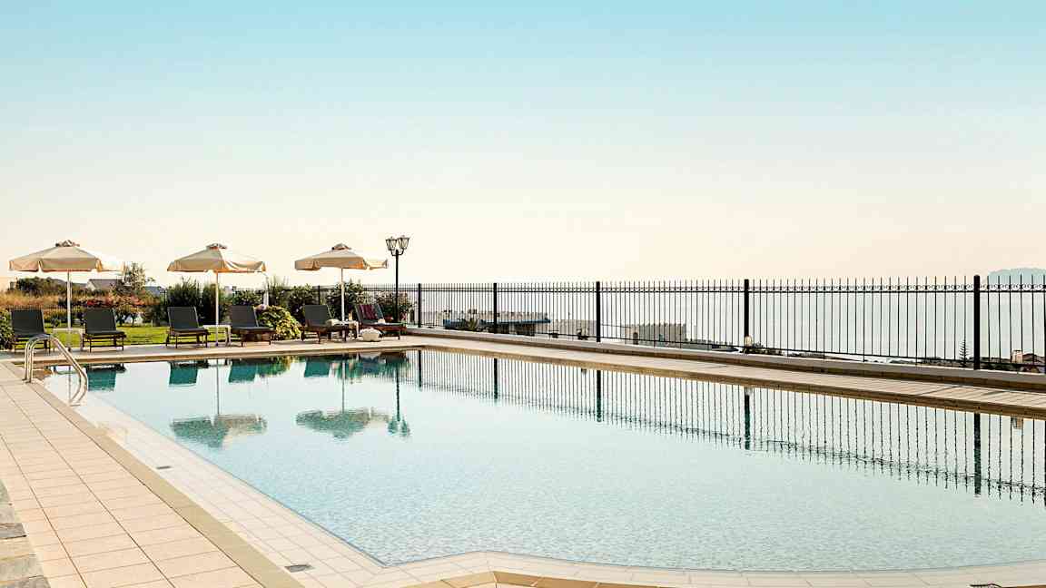 Et utendørs svømmebasseng med solsenger og parasoller ved siden av, med havet i bakgrunnen. (AI-generert tekst)