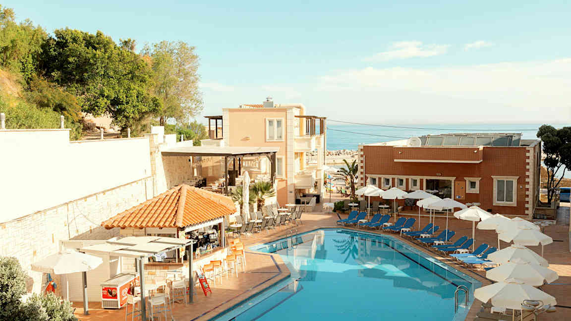 Et hotellbasseng med solsenger og parasoller ved siden av, samt bygninger og grøntområder i bakgrunnen. (AI-generert tekst)