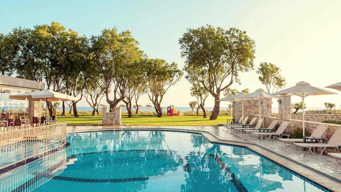 Et basseng med solsenger og parasoller omgitt av grøntområder og trær. (AI-generert tekst)