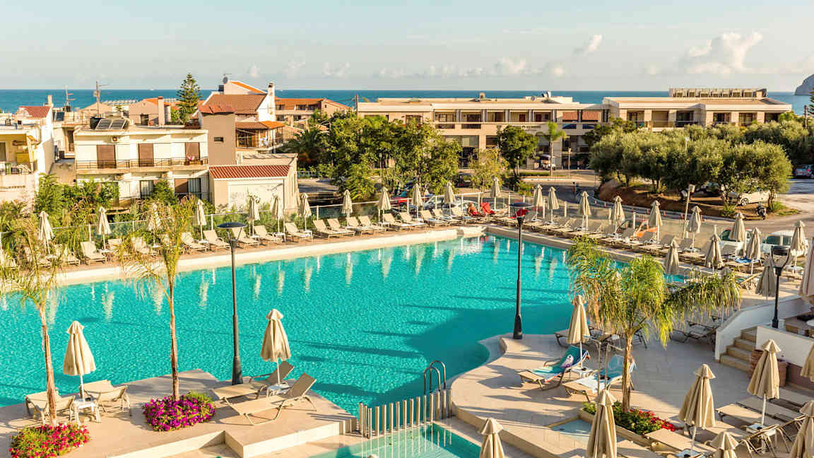 Et utendørs svømmebasseng omgitt av solsenger og parasoller, med bygninger og havet i bakgrunnen. (AI-generert tekst)