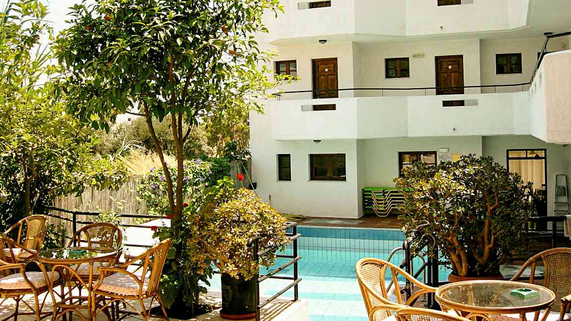 Et utendørs basseng omgitt av vegetasjon og sittemøbler, samt en hvit bygning med balkonger i bakgrunnen. (AI-generert tekst)