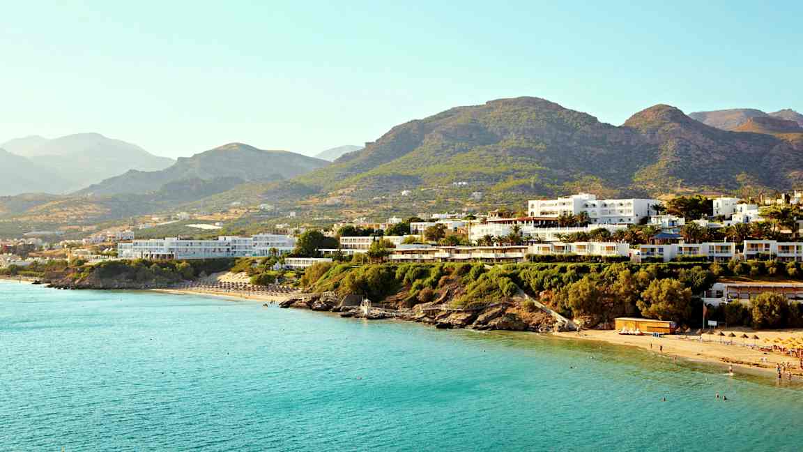 Flere bygninger nær en kyst med blått hav og fjell i bakgrunnen. (AI-generert tekst)