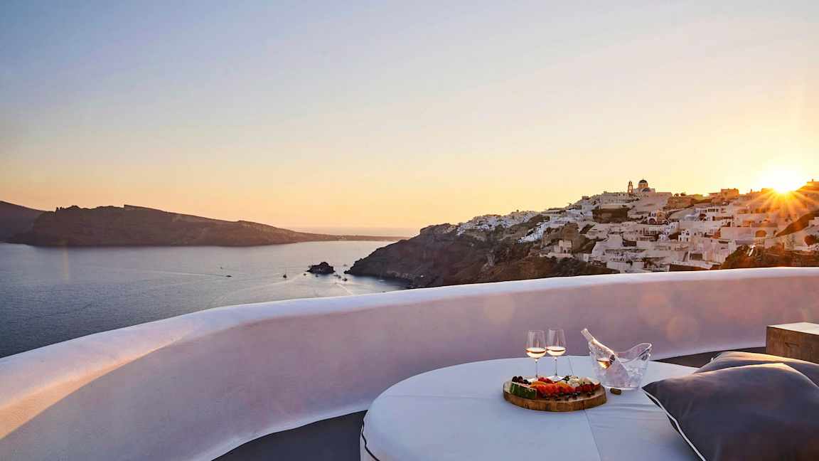 Utsikt over en solnedgang ved sjøen med hvite bygninger på en klippe og en terrasse med bord, mat og drikke i forgrunnen. (AI-generert tekst)