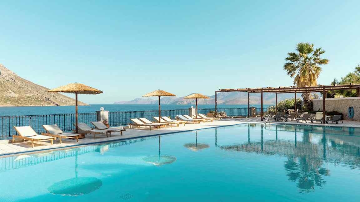 Et utendørs svømmebasseng med solsenger og parasoller ved siden, samt havutsikt i bakgrunnen. (AI-generert tekst)