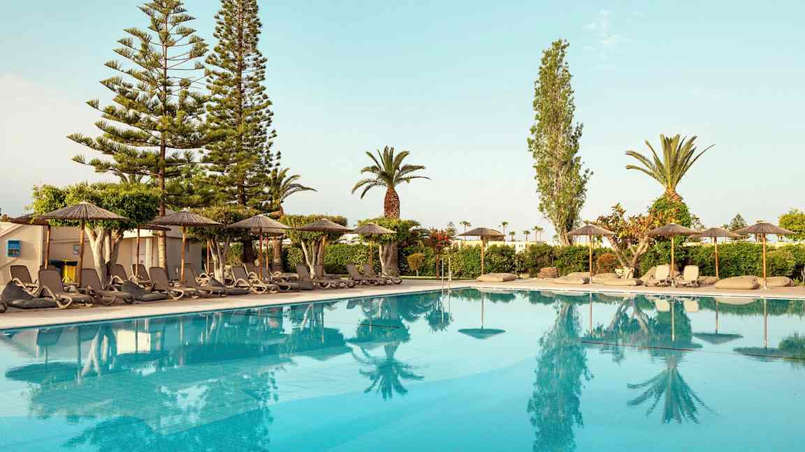 Utsikt over et basseng med solsenger og parasoller på kanten, omgitt av trær og busker. (AI-generert tekst)