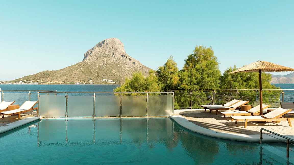 Et utendørsbasseng med solsenger og parasoll, plassert nær havet med en ås i bakgrunnen. (AI-generert tekst)