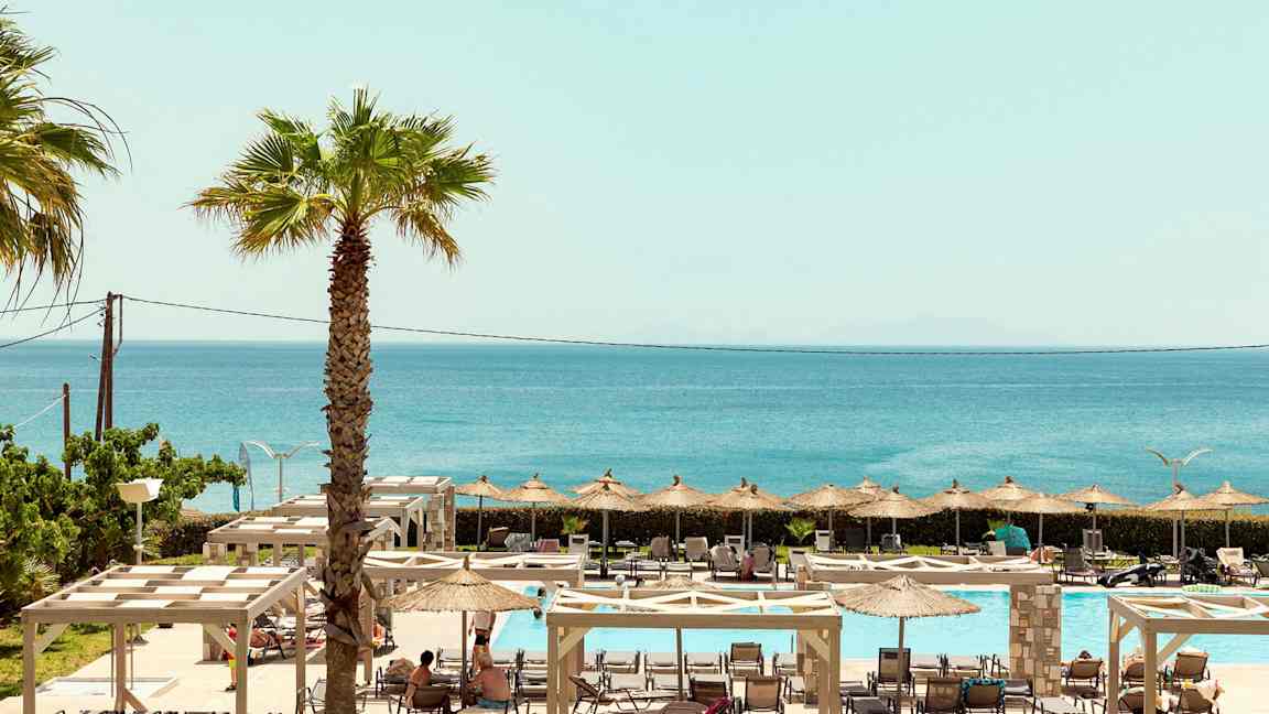 En utsikt over utendørs bassengområde med solsenger, parasoller og havet i bakgrunnen. (AI-generert tekst)