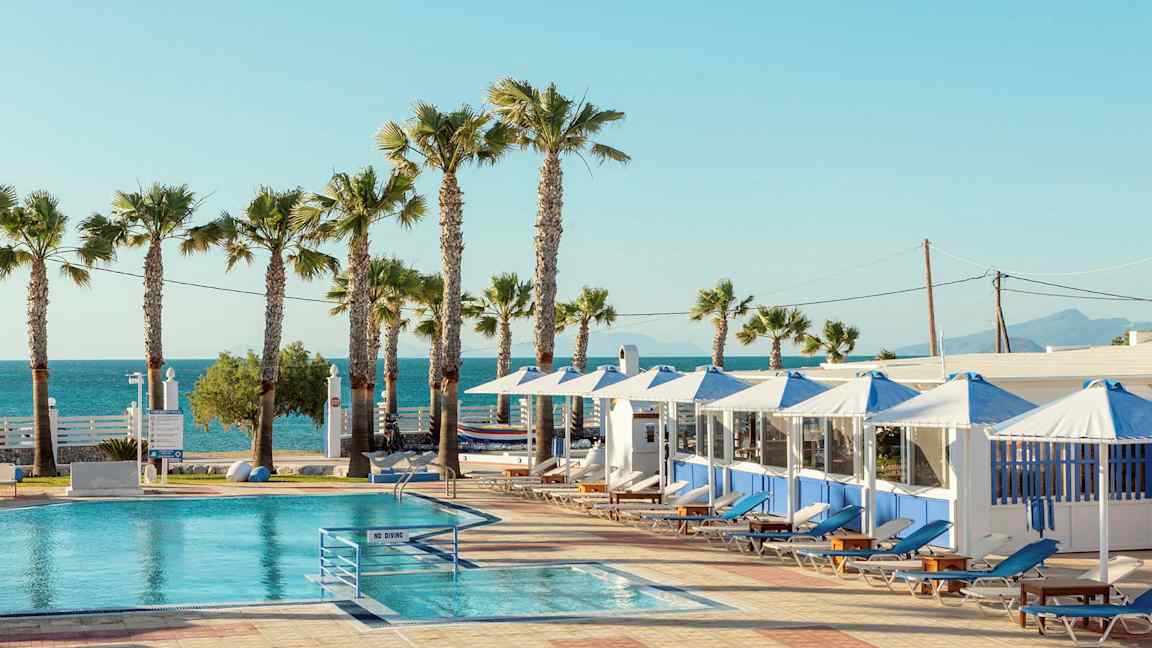 Et basseng med solsenger og parasoller under en klar himmel. Palmer og hav i bakgrunnen. (AI-generert tekst)