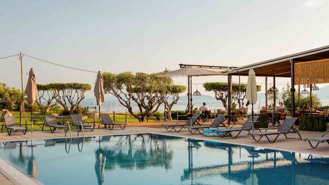 Utsikt over et basseng med solsenger rundt omkring, trær i bakgrunnen og havet i det fjerne. (AI-generert tekst)