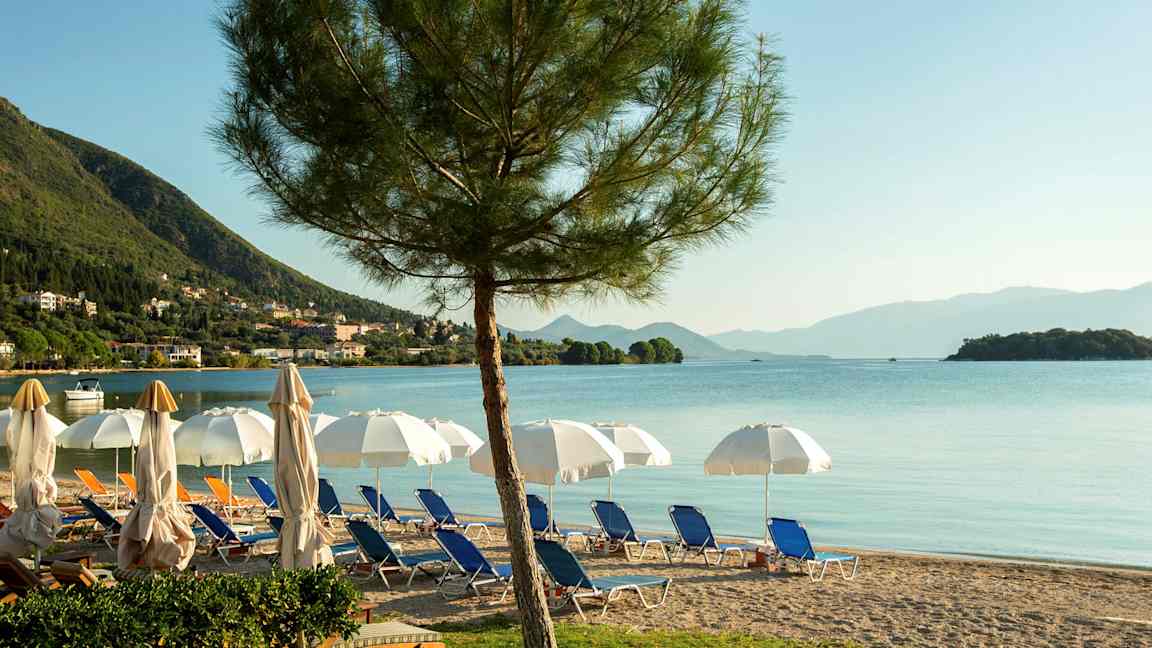 En strand med solsenger, parasoller og utsikt over havet med fjell i bakgrunnen. (AI-generert tekst)