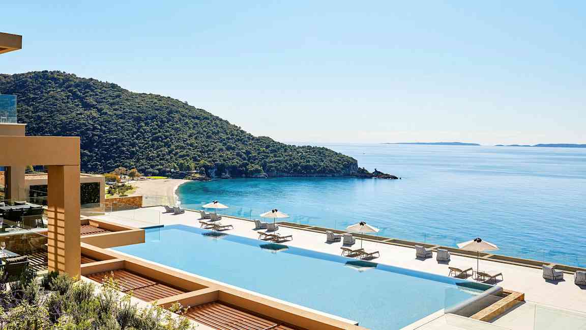 Utsikt over et basseng med solsenger og parasoller, en kystlinje og hav i bakgrunnen. (AI-generert tekst)