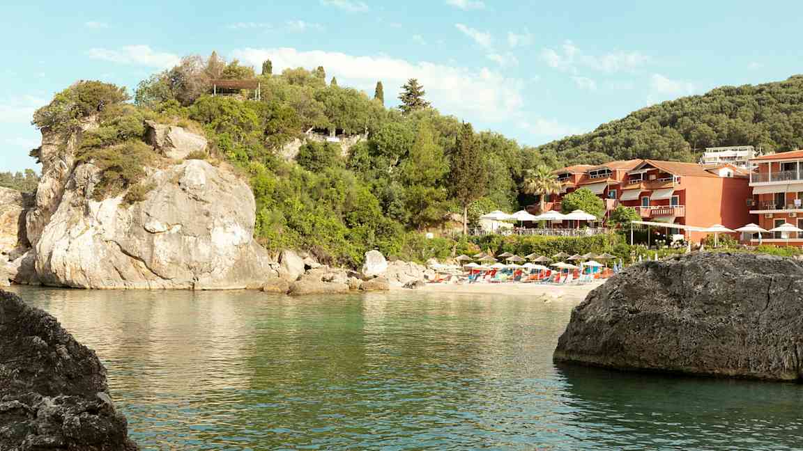 Bilde av en klippestrand med parasoller og solsenger samt bygninger omgitt av grøntområder. (AI-generert tekst)