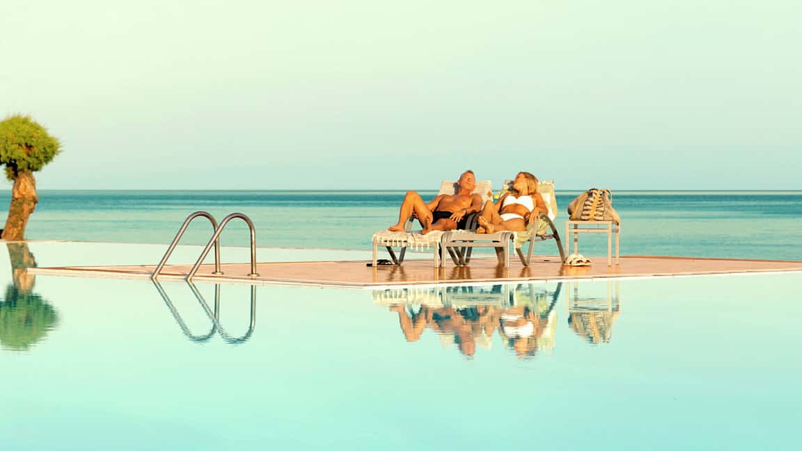 To personer ligger på solsenger ved et basseng med utsikt over havet. (AI-generert tekst)