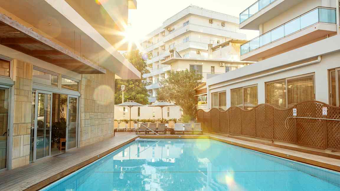 Utendørs svømmebasseng med omkringliggende solsenger og bygninger. (AI-generert tekst)