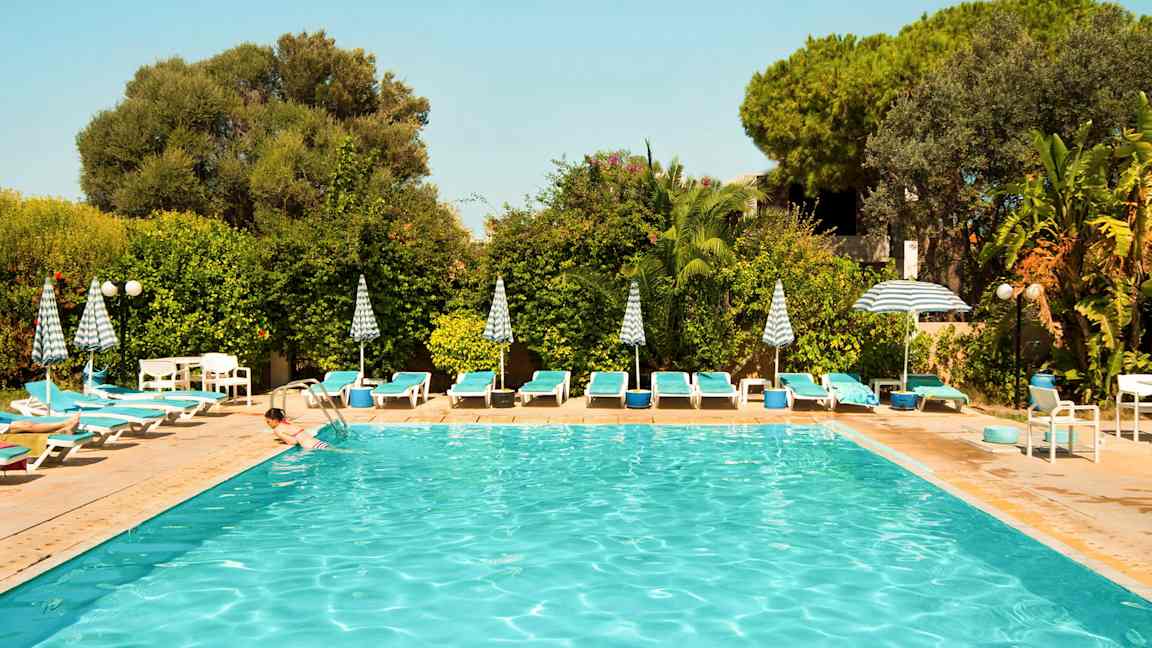 Utendørs svømmebasseng omgitt av solsenger, parasoller og grønne planter. (AI-generert tekst)