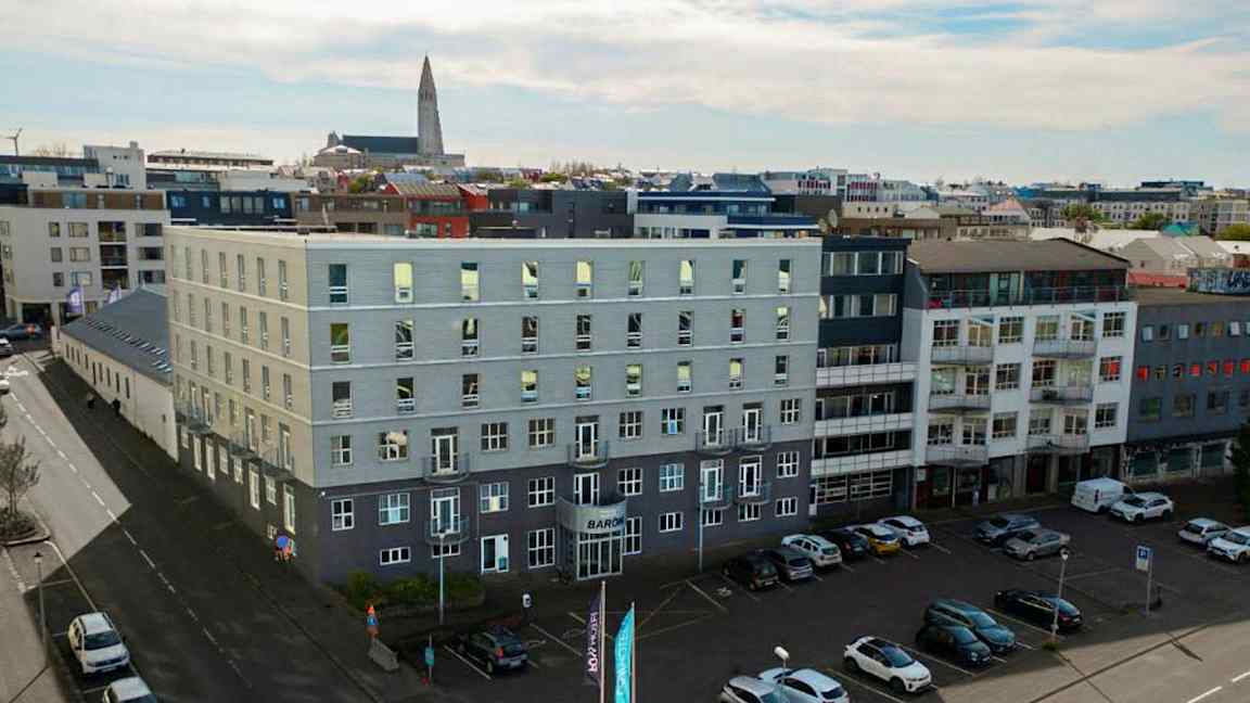 Flere moderne bygninger omgir et åpent parkeringsområde, med biler parkert på bakken og en kirke synlig i bakgrunnen. (AI-generert tekst)