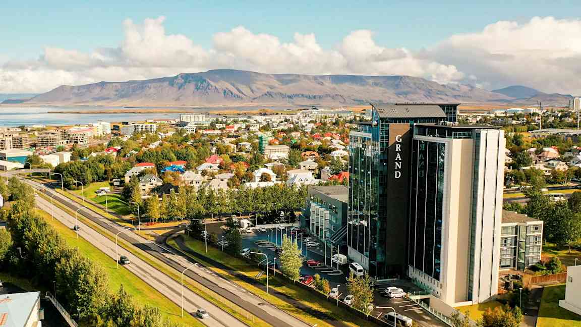 Et bymiljø med en motorvei i forgrunnen, trær, bygninger og et større hotell. I bakgrunnen sees et fjellandskap. (AI-generert tekst)