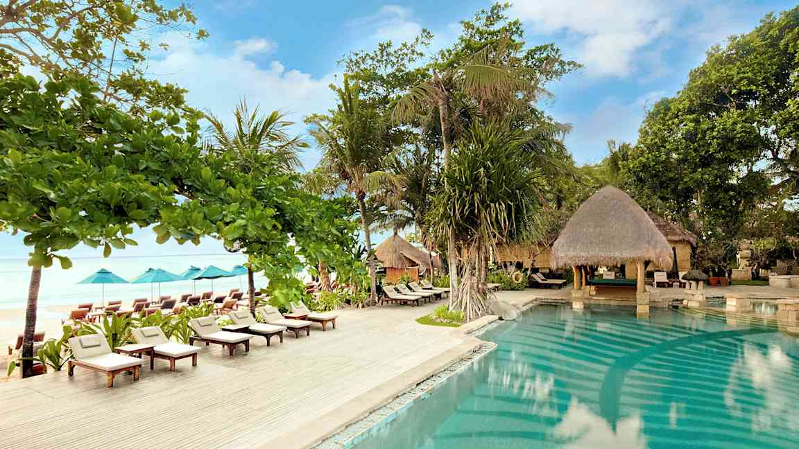 Et utendørs svømmebasseng omgitt av solsenger, parasoller og natur ved sjøen. (AI-generert tekst)