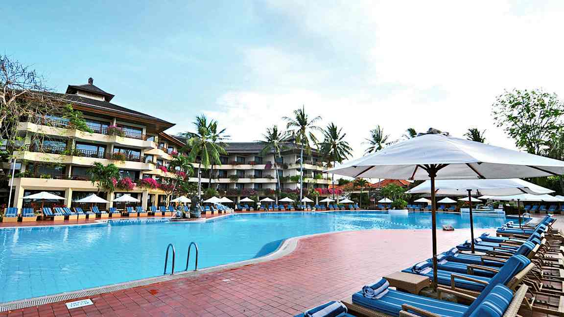 Bilde av et utendørs svømmebasseng omgitt av solsenger og parasoller, med et fleretasjes hotell og palmer i bakgrunnen. (AI-generert tekst)