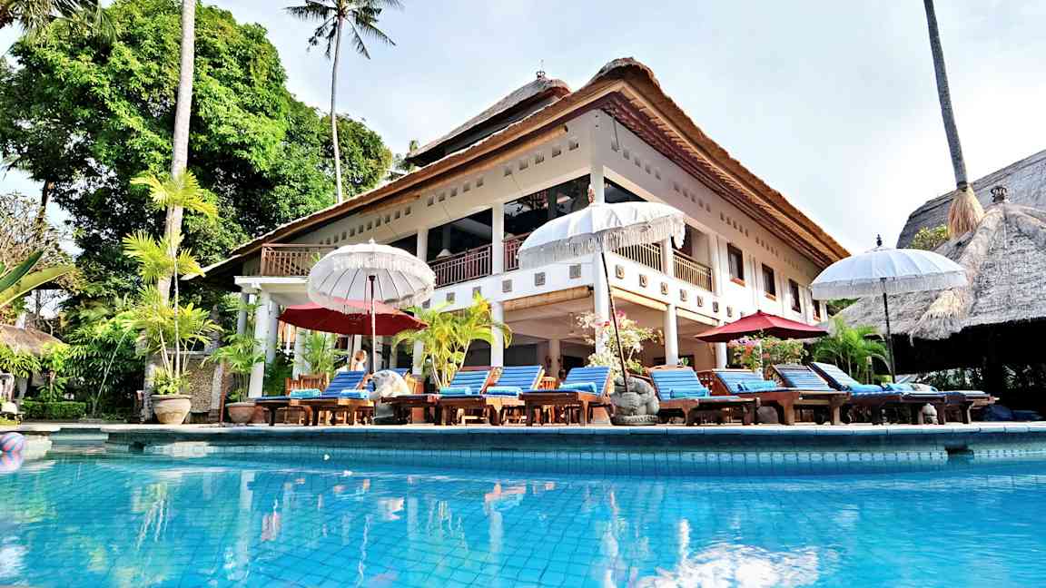 En stor villa med stråtak og et basseng foran, omgitt av solsenger og parasoller. (AI-generert tekst)