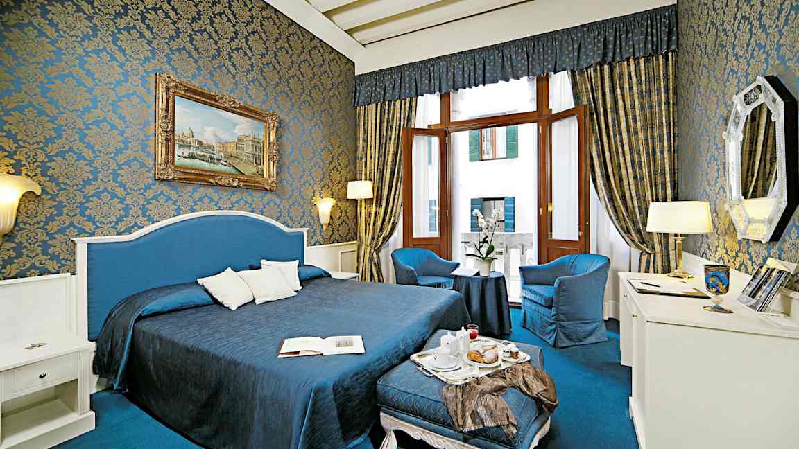 Et vakkert innredet hotellrom med blå og gyldne detaljer, stor seng, sittegruppe og store vinduer. (AI-generert tekst)