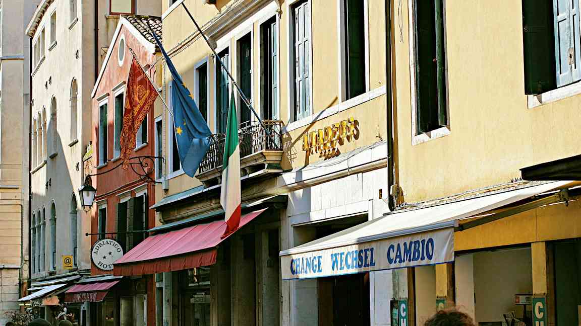 Fasade av bygninger med ett italiensk flagg, europeisk flagg og skilt som angir hotell og veksling. (AI-generert tekst)