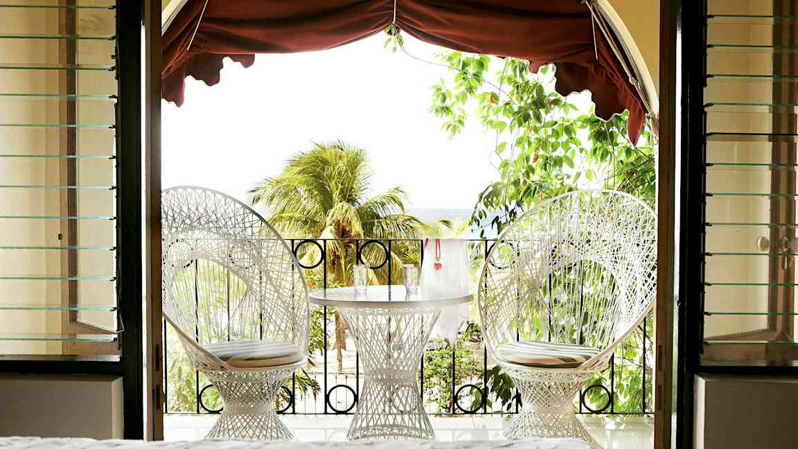 Utsikt fra et rom mot en terrasse med to hvite stoler og et bord med glass, omgitt av grønt og palmer. (AI-generert tekst)