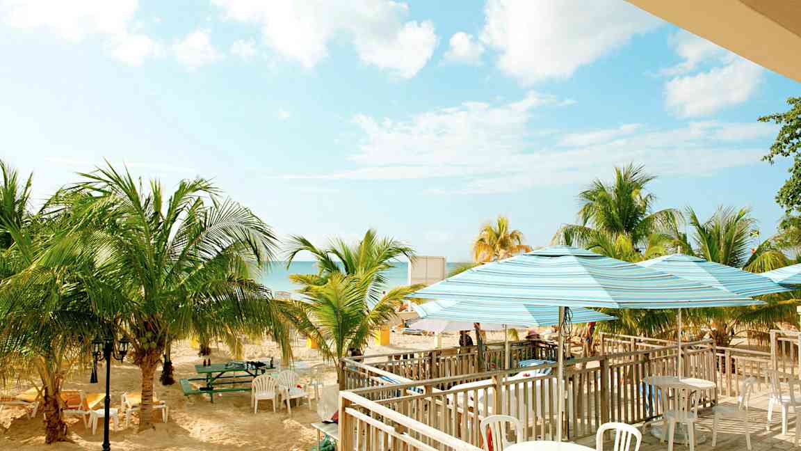 Bilde av en strand med palmer, bord og stoler samt parasoller. Himmelen er klar med noen skyer. (AI-generert tekst)