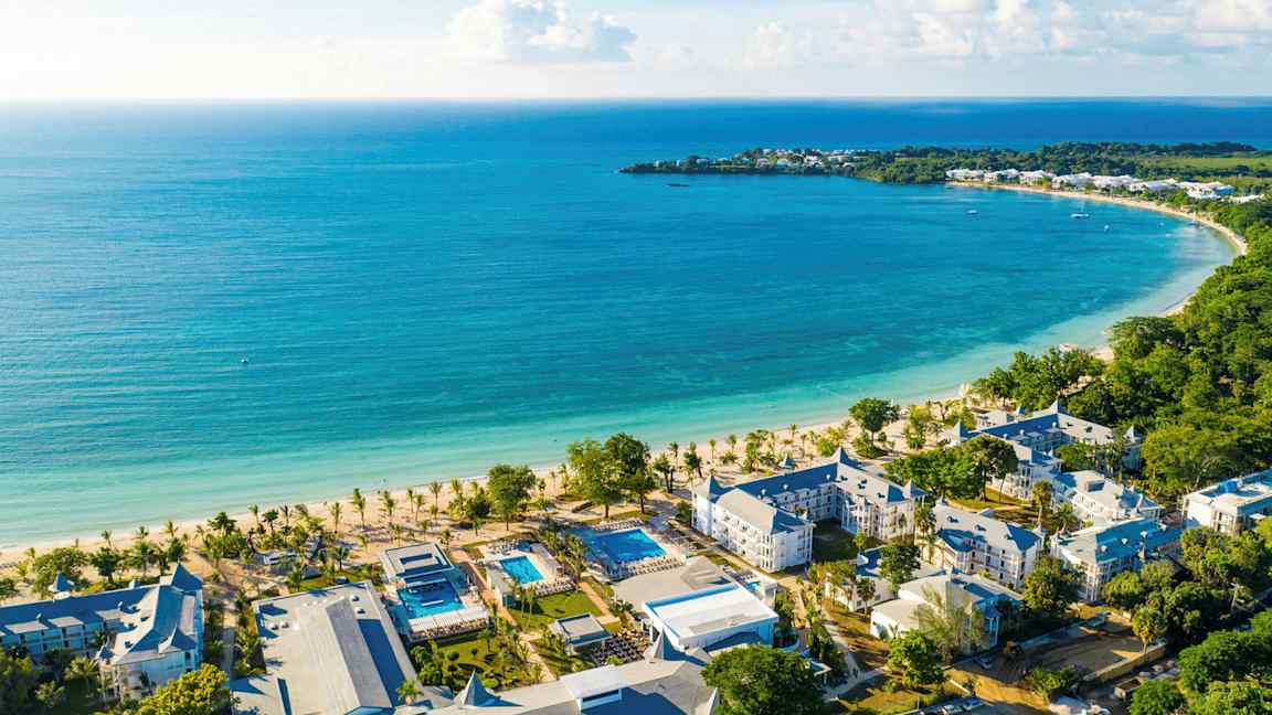 Bilde av en kyststrekning med turkis vann, en hvit sandstrand og hotellbygninger i nærheten. (AI-generert tekst)
