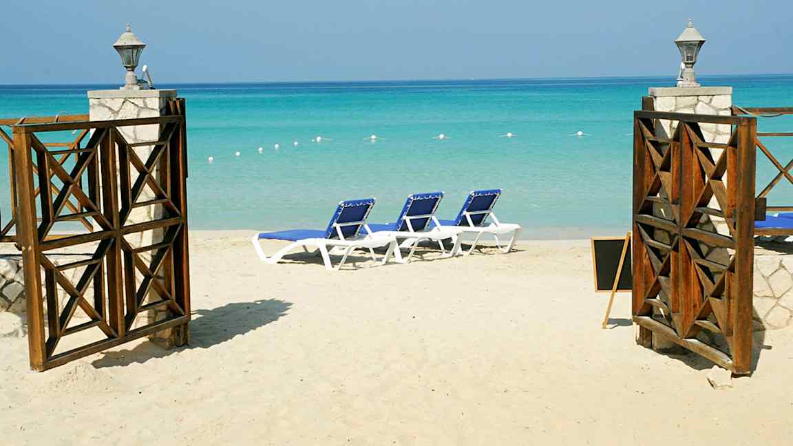 En sandstrand med solsenger og en åpen port med utsikt over det turkise havet. (AI-generert tekst)