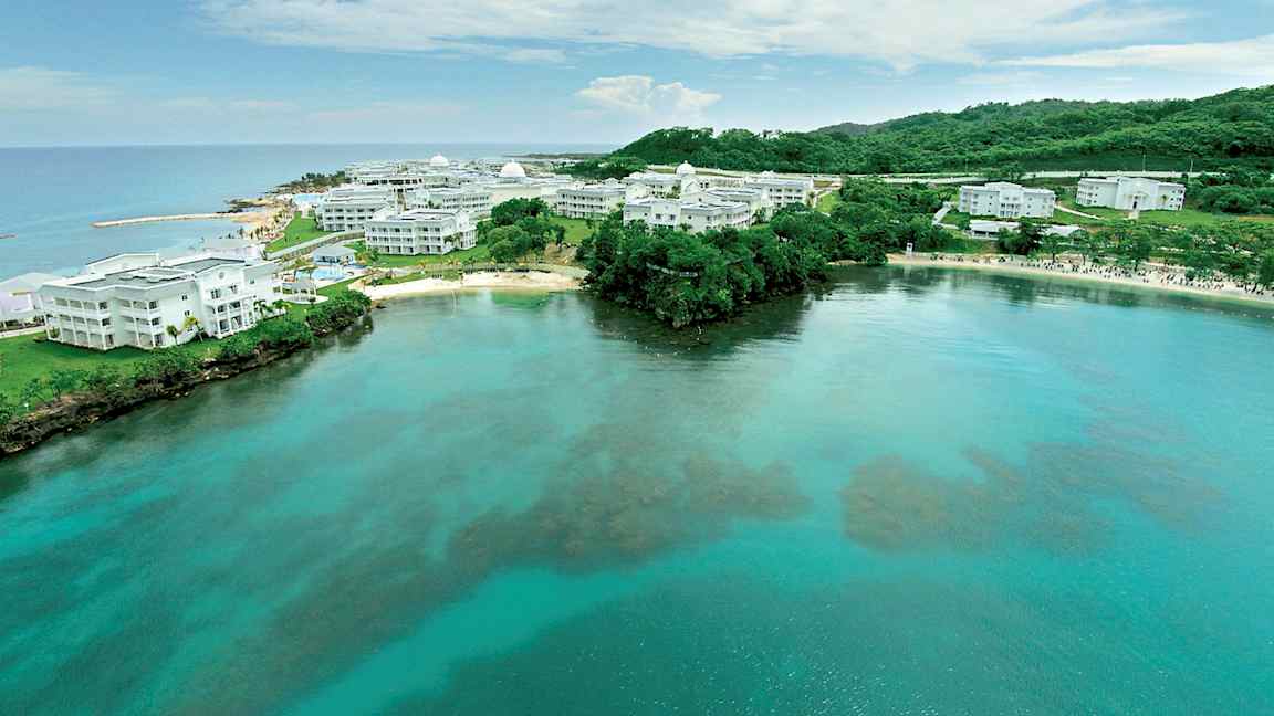 Bilde av en kyst med klart vann og hvite bygninger omgitt av grøntområder. (AI-generert tekst)