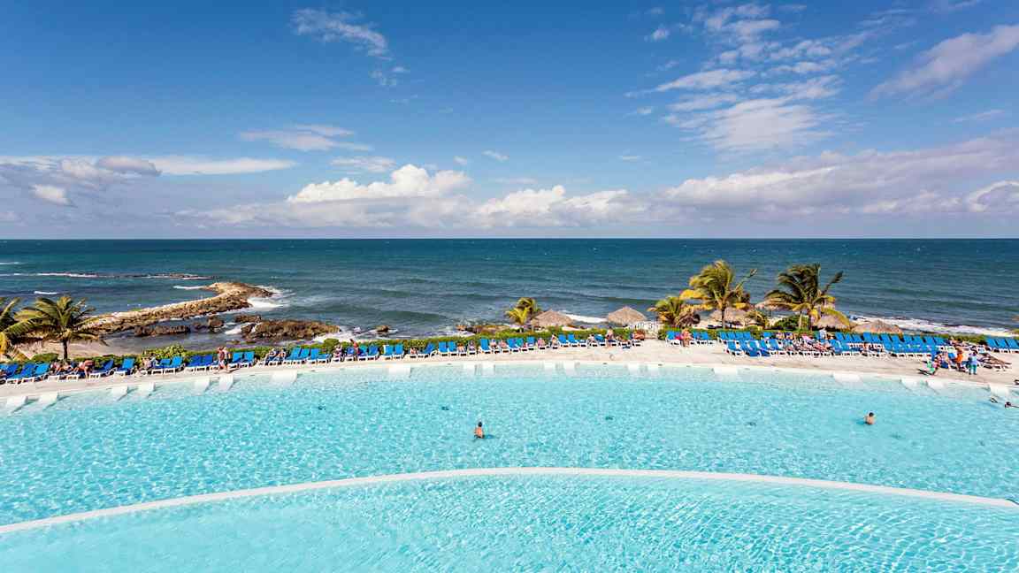 Stor basseng foran havet med solsenger og palmer. (AI-generert tekst)