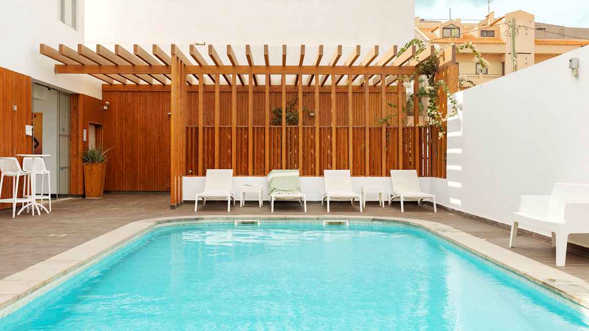 Et lite basseng med omgivende solsenger og treterrasse, en treramme skaper en avskjermet atmosfære. (AI-generert tekst)