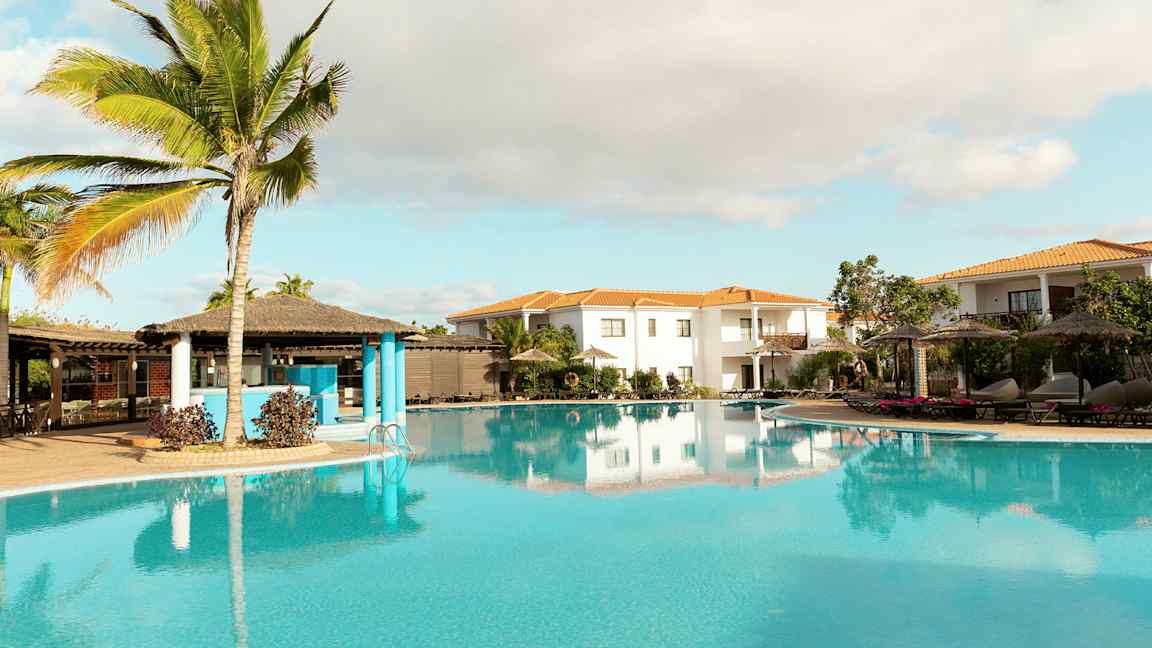 En tropisk bassengområde med palmer, parasoller og omkringliggende bygninger. (AI-generert tekst)