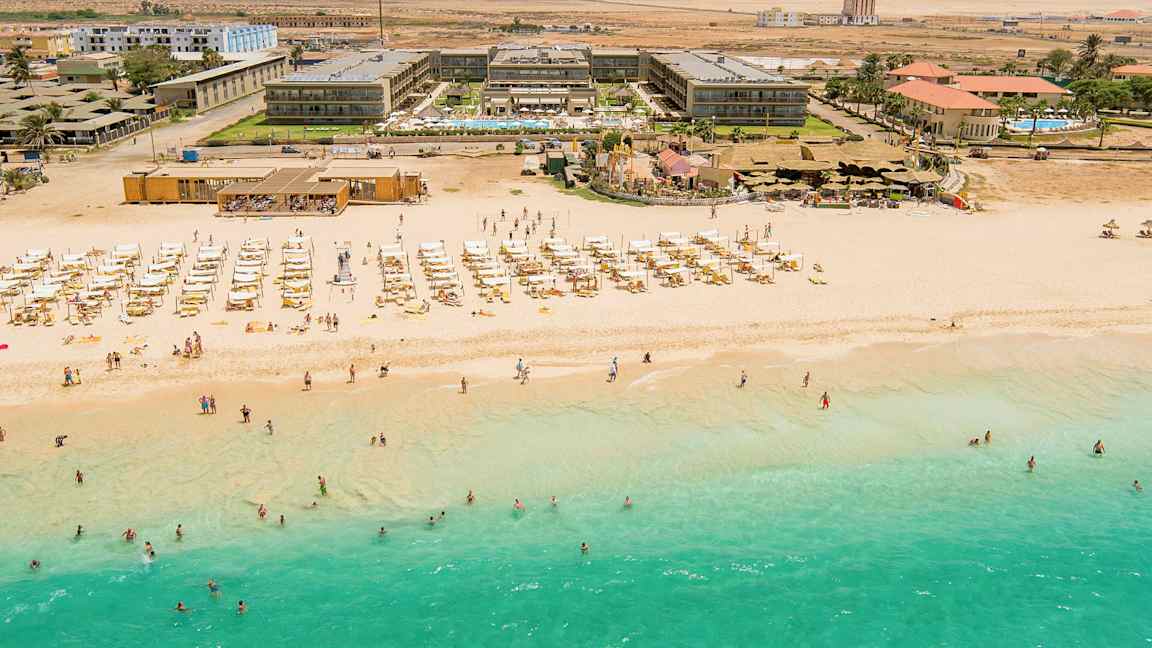 Luftfoto av en strand med turkis vann, solsenger og parasoller, samt bak en hotellbygning. (AI-generert tekst)