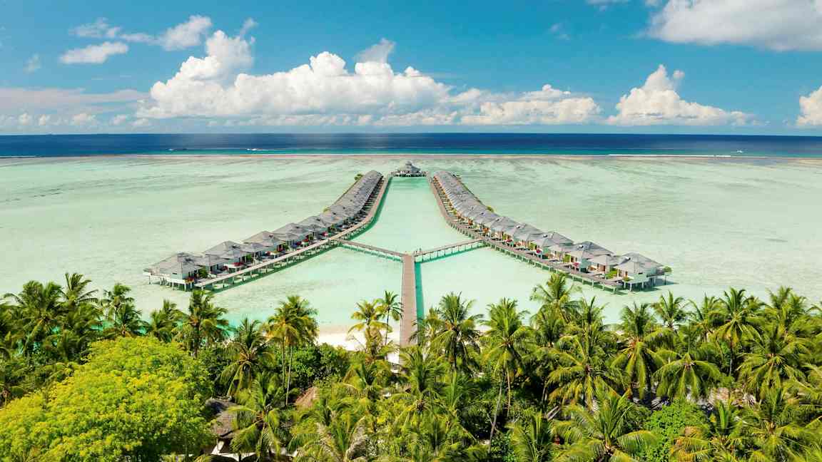 Bildet viser over-vanns bungalower koblet sammen med brygger over en lagune, omgitt av palmer og hav med en blå himmel. (AI-generert tekst)