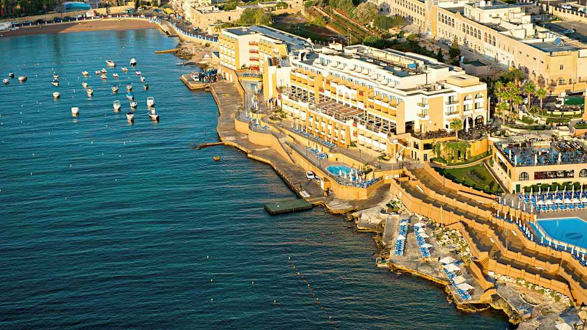 Luftbilde av et kysthotell med bassenger og solsenger ved sjøen. Små båter er fortøyd i vannet. (AI-generert tekst)