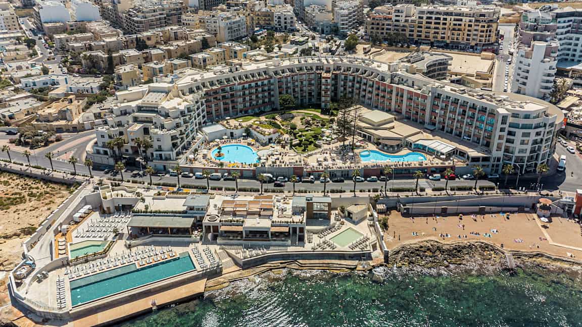 Bildet viser en omfattende ferieanlegg med bassenger, solområder og omkringliggende bygninger nær kysten. (AI-generert tekst)