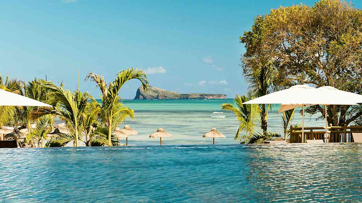 Et infinitybasseng med utsikt over en strand med parasoller og en liten øy i havet. (AI-generert tekst)