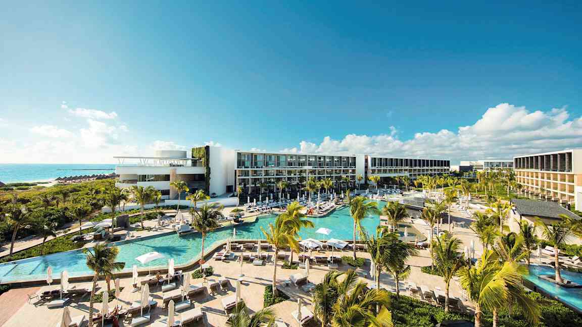 Et moderne hotellkompleks med flere bassenger, frodig vegetasjon og palmer, beliggende ved kysten med utsikt over havet under en klar blå himmel. (AI-generert tekst)