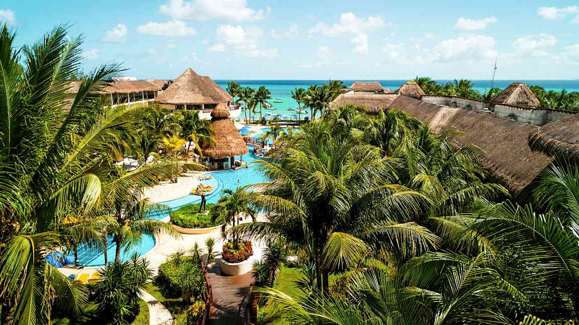 Et stort feriested med bassenger, palmer og bygninger med stråtak, plassert nær havet under en blå himmel. (AI-generert tekst)