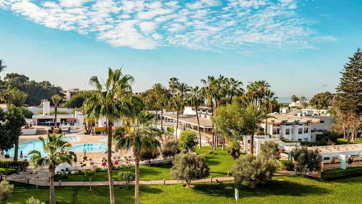 Hotellområde med gressplener, palmer, basseng og hvite bygninger. (AI-generert tekst)