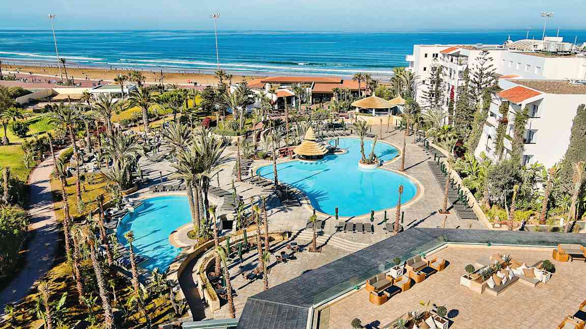 Hotellområde med bassenger og omkringliggende hager, plassert nær en sandstrand og havet. (AI-generert tekst)