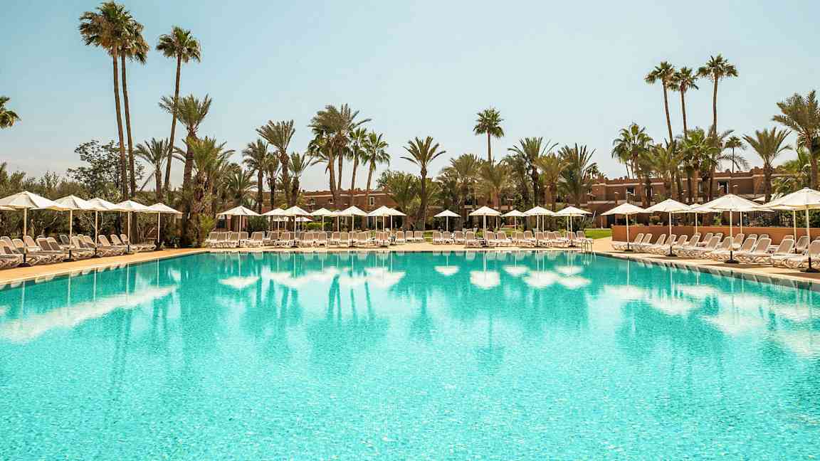 Et stort basseng omgitt av solsenger og parasoller under en klar himmel med palmer i bakgrunnen. (AI-generert tekst)