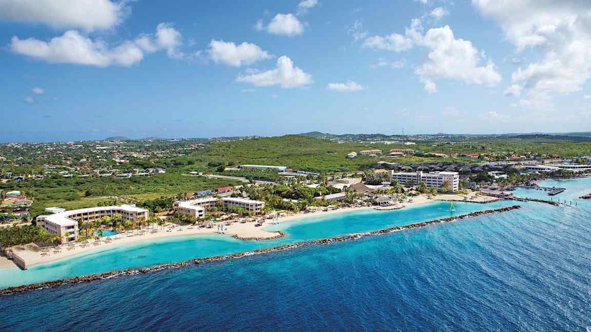 Luftfoto av en kystlinje med hotellbygninger ved en strand og grøntområder i bakgrunnen. (AI-generert tekst)
