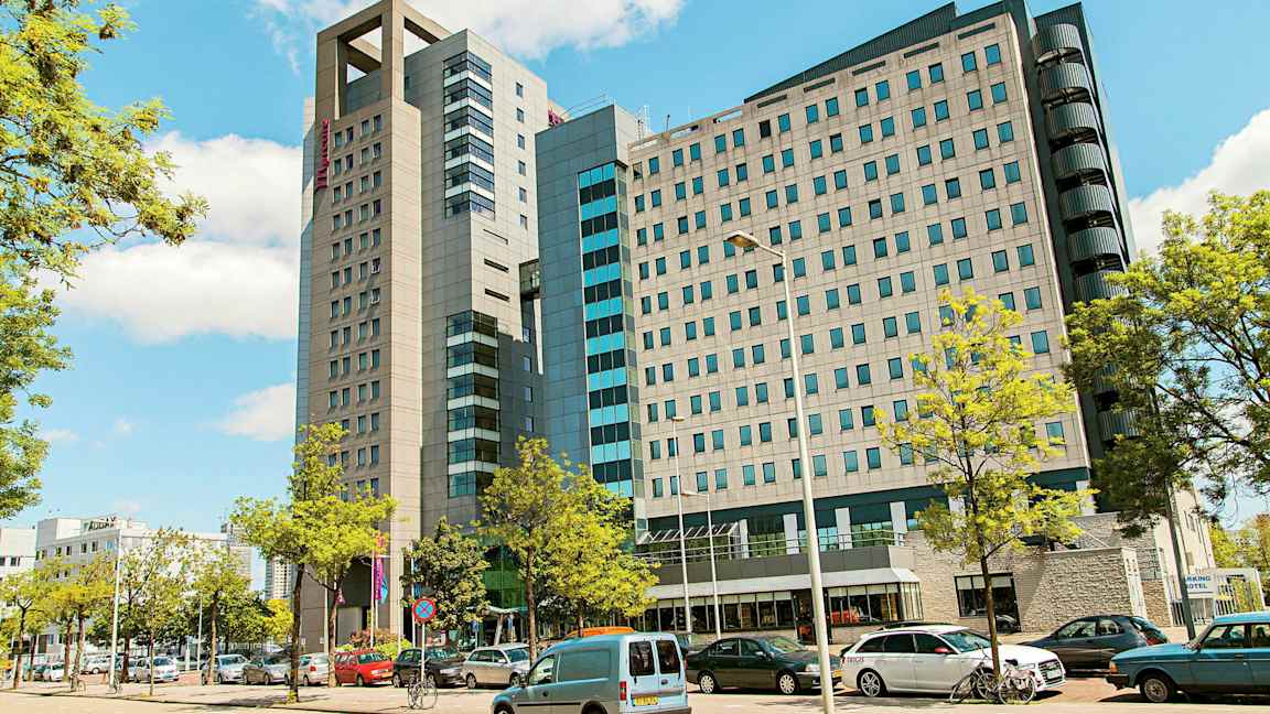 En moderne bygning med flere etasjer omgitt av parkerte biler og trær. (AI-generert tekst)